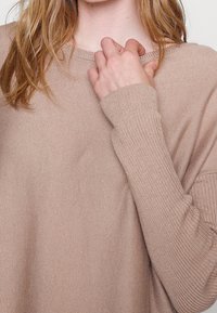 Frau mit langen Haaren, die einen beigen gestrickten Pullover trägt und den Halsausschnitt mit ihrer rechten Hand hält.