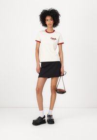 Crème shirt met korte mouwen en een rode geribde kraag en manchetten, met het "Coach New York" logo. Gecombineerd met een zwarte mini rok en platform schoenen.