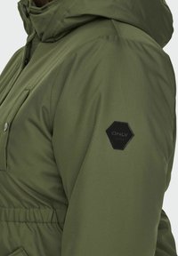 Olivgrön puffjacka med huva, framficka och en svart hexagonal logotyp på ärmen. Slät, strukturerad tyg.