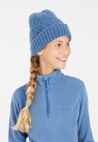 Berretto lavorato a maglia blu con un motivo testurizzato e un piccolo logo in legno marrone. Abbinato a un top in pile azzurro chiaro con colletto con zip.