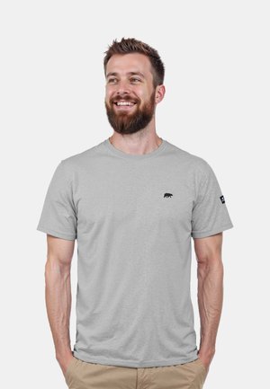 Camiseta de color gris claro en una tela suave, con un pequeño logotipo de oso negro en el pecho y una etiqueta en la manga. Diseño de mangas cortas y cuello redondo.