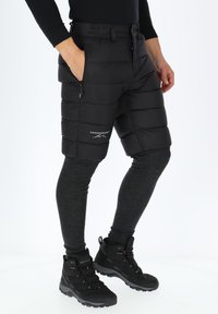 Zwarte geïsoleerde shorts met een horizontaal gewatteerd patroon, ritszakken en logo. Draagt over donkergrijze leggings en gecombineerd met zwarte wandelschoenen.