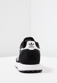Mustat ja valkoiset Adidas-tennarit nähtynä takaa, jossa on Adidas-logo kantapään lenkissä ja teksturoitu valkoinen pohja.