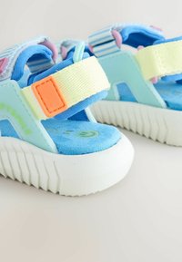 Sandales pour enfants bleu et vert pastel avec semelles blanches épaisses, sangles jaunes et détail de boucle orange, posées sur une surface claire.