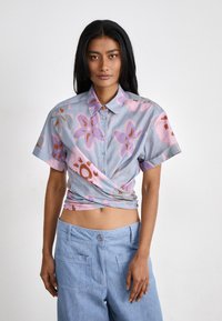 Camicia a maniche corte, con bottoni, di colore azzurro chiaro, decorata con motivi floreali rosa e arancioni, caratterizzata da un design annodato in vita. Tessuto morbido.