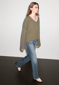 Olive green cable-knit sweater with a V-neck, worn over a white top, paired with flared blue jeans and open-toe heels. Simple design.