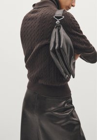 Borsa a tracolla in pelle nera con design a pieghe, agganciata da accessori in metallo, indossata sopra un maglione marrone a maglia cable e una gonna in pelle liscia.