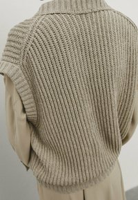 Beige strikket vest med ribbet tekstur, korte ærmer og høj krave, med en løs pasform og rundet kant.