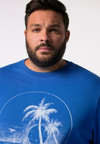 Modré bavlněné tričko s grafickým potiskem palem na bílém kruhovém pozadí slunce. Kulatý výstřih a krátké rukávy.