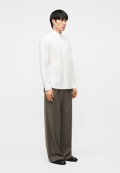 Νεαρός άνδρας όρθιος, φοράει λευκό πουκάμισο με κουμπιά, φαρδιά παντελόνια σε σκούρο καφέ χρώμα και μαύρα παπούτσια, μπροστά σε απλό λευκό φόντο.