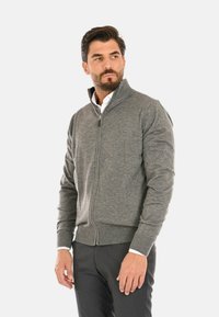 Cardigan grigio con zip e colletto alto, texture a maglia e polsini a coste. Presenta una zip liscia e un design aderente.
