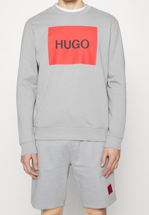 Męski model noszący jasnoszary zestaw bluzy i szortów, bluza ma duży czerwony kwadrat z napisem "HUGO" na piersi.