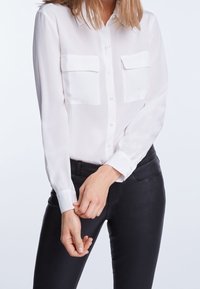 Femme en chemise blanche à boutons avec poches poitrine ajustant le poignet, portant un pantalon noir ajusté sur un fond uni.