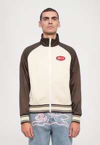 Bruine en crèmekleurige zip-up jas met hoge kraag en geribde zoom, met een rode patch op de borst. Getextureerde stof met contrasterende mouwen.