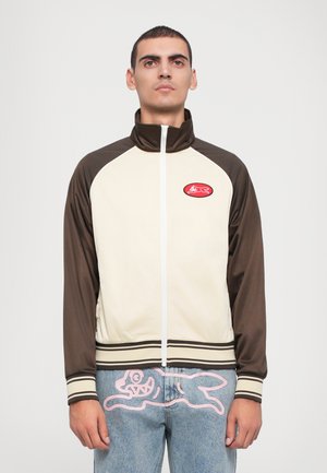 Bruine en crèmekleurige zip-up jas met hoge kraag en geribde zoom, met een rode patch op de borst. Getextureerde stof met contrasterende mouwen.