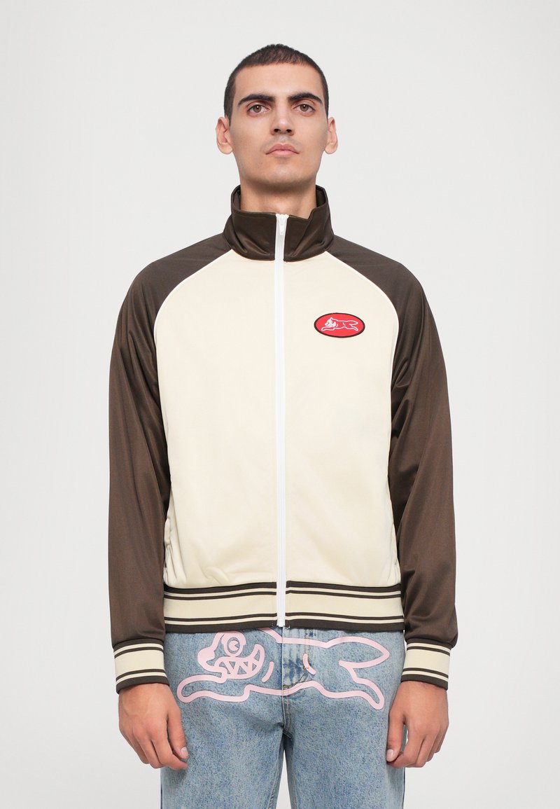 Bruine en crèmekleurige zip-up jas met hoge kraag en geribde zoom, met een rode patch op de borst. Getextureerde stof met contrasterende mouwen.