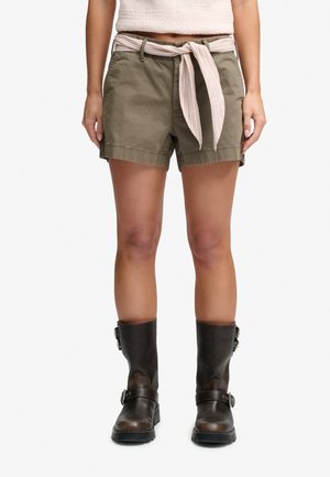 Persoon draagt olijfgroene korte broek met een licht gestreepte stoffen riem gestrikt in de taille en donkerbruine halfhoge laarzen met gespen.