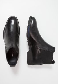 Musta nahkasaapas nilkkamitoilla, jossa on joustavat sivupaneelit, pyöristetyt varpaat ja karkea kumipohja, jossa on vaakaura ja sinisiä yksityiskohtia.