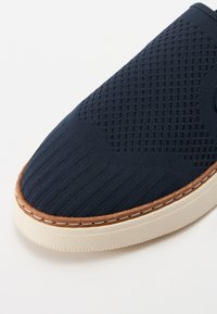 Tamno plava slip-on cipela s teksturiranim mrežastim gornjim dijelom, opečastim kožnim detaljima, svjetlosnim gumеним potplatom i suptilnim šavnim detaljima.