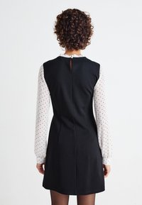 Mustapuku, jossa on tiivis yläosa, levenevä hame ja avainaukko selässä. Pitkissä valkoisissa hihoissa on mustia pilkkuja ja röyhelökaulus.