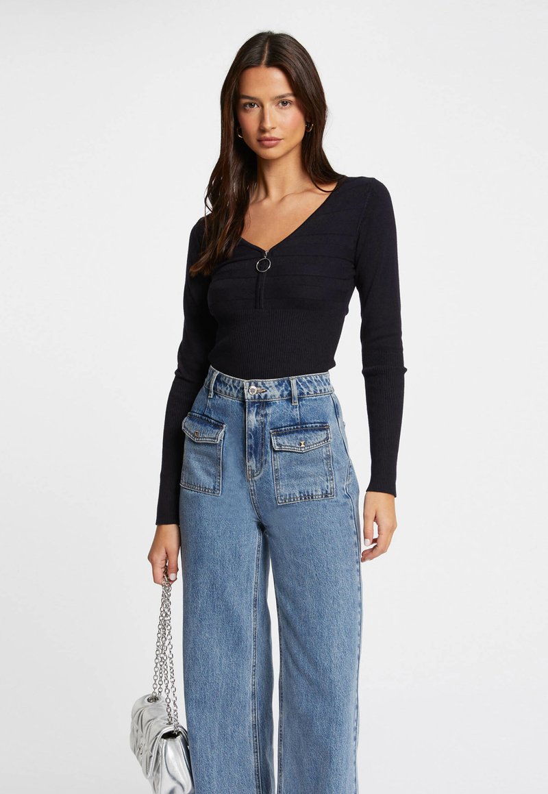 Schwarzes, geripptes Langarmoberteil mit V-Ausschnitt und einem runden Reißverschluss-Detail, kombiniert mit hochgeschnittenen, hellblauen weiten Jeans und einer silbernen Handtasche.