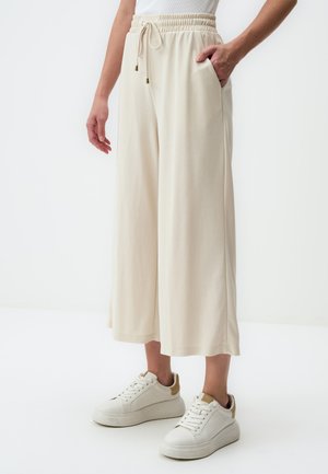 Pantaloni larghi color crema con vita elastica e dettagli con cordino. Presentano tasche laterali e una texture morbida e liscia. Abbinati a sneakers bianche.