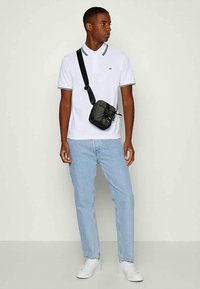 Νεαρός άνδρας φορώντας λευκό πόλο, ανοιχτό μπλε τζιν, λευκά αθλητικά παπούτσια και μια μικρή μαύρη τσάντα χιαστί, στέκεται και κοιτάζει δεξιά.