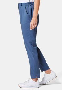Pantalons bleu extensibles avec une taille élastique, des plis à l'avant, des poches latérales et des fentes à l'ourlet, associés à des baskets blanches.