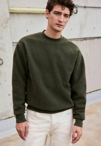 Olivengrüner Fleece-Pullover mit Rundhalsausschnitt und gerippten Bündchen. Bietet einen lässigen Schnitt und eine weiche Textur, kombiniert mit hellbeigen Hosen.