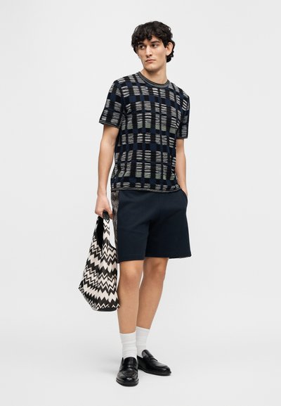 Μπλε ναυτικό μπλουζάκι με οριζόντιες ρίγες, σε συνδυασμό με σκούρο σορτς. Κρατάει μια μαύρη και λευκή τσάντα tote με ζιγκ-ζαγκ μοτίβο. Μαύρα παπούτσια τύπου loafers και λευκές κάλτσες.