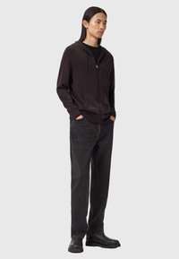 Ung man med långt hår iförd en mörk zip-up hoodie, svart t-shirt, urblekta svarta jeans och svarta kängor som står med händerna i fickorna.