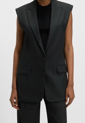 Blazer noir sans manches avec épaules rembourrées porté sur un haut côtelé noir, assorti à un pantalon noir sur mesure.