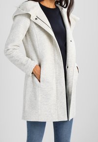 Abrigo con capucha color gris claro, fabricado con tejido texturizado, con un cierre de broche singular y bolsillos laterales. Lleva puesto sobre una prenda oscura y combinado con unos vaqueros azules.