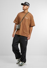 Brun oversized t-shirt med logo, sorte bukser med detaljer, grønne og hvide sneakers, samt en lille crossbody taske i en afslappet stående pose.