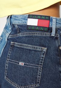 Blå denimjeans med hög midja, med en svart lapp märkt "TOMMY JEANS" med en röd, vit och grön logotyp på bakre midjebandet.