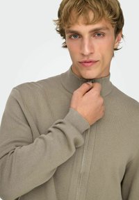 Giacca beige con cerniera, collo alto, tessuto lavorato a maglia e polsini a coste. Modello che tiene la cerniera, mostrando i dettagli.