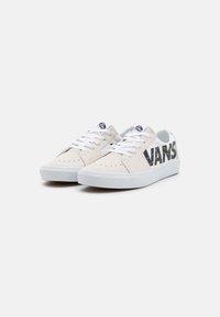 Vita kanvas sneakers med svart "VANS"-logotyp, vita snören, perforerad detaljering på tån och en vit gummisula.