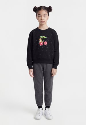 Ragazza con doppio chignon che indossa una felpa nera con motivo di ciliegie e orsacchiotto, pantaloni da jogging grigi e sneakers bianche, in piedi contro uno sfondo bianco.