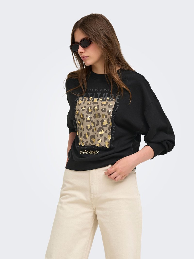 Femme portant des lunettes de soleil noires, un pull noir avec imprimé léopard doré et texte, et un pantalon beige clair sur un fond clair uni.