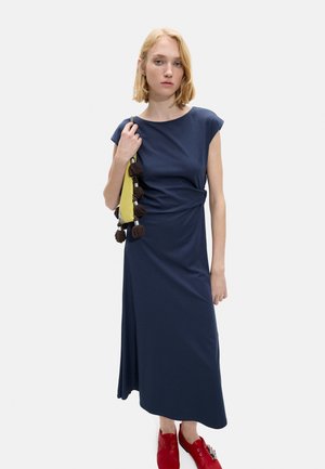 Kobieta o blond włosach, ubrana w granatową sukienkę, trzyma żółtą torebkę z brązowymi frędzlami i nosi czerwone buty.