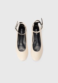 Une paire de chaussures plates beiges avec des brides de cheville et des semelles noires, photographiée de dessus sur un fond blanc.