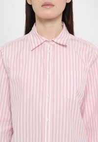 Femme portant une chemise rayée verticalement rose et blanche avec un col et une poche poitrine, montrée du menton à la taille.