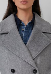 Grijze wollen peacoat met een dubbelgevouwen ontwerp, zwarte knopen en een stevige kraag, gedragen over een spijkerhemd.