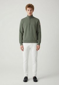 Oliivinvihreä vetoketjullinen collegepaita, jossa on korkea kaulus, resorit hihansuissa ja helmassa, yhdistettynä valkoisiin kapeisiin housuihin ja mustiin loafer-tyylisiin kenkiin.