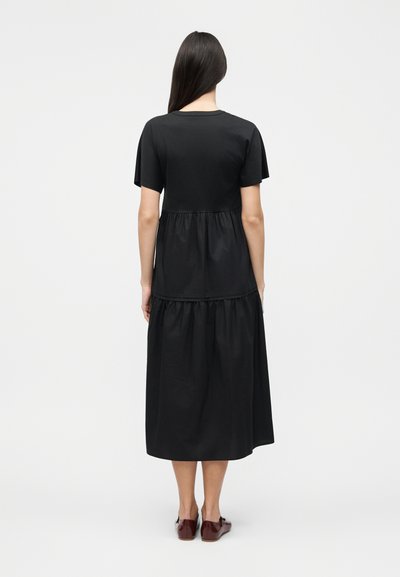 Γυναίκα που στέκεται με την πλάτη, φοράει μαύρο midi φόρεμα με κοντά μανίκια και καφέ επίπεδα παπούτσια ενώ ποζάρει σε απλό λευκό φόντο.