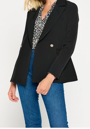 Vrouw draagt een zwart dubbelgeknoopt colbert over een zwart-wit patroon blouse en blauwe spijkerbroek, met een dunne gouden armband om haar pols.
