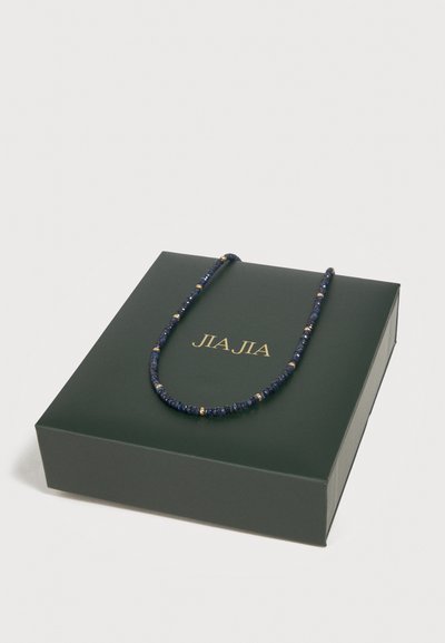 Σκουρό μπλε κολιέ με χάντρες και χρυσές λεπτομέρειες, τοποθετημένο σε σκούρο πράσινο κουτί με χρυσό κείμενο «JIA JIA» σε λευκό φόντο.