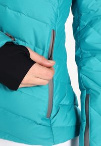 Turkusowa pikowana kurtka z zamykanymi na zamek kieszeniami. Posiada gładką teksturę i dopasowany krój. Czarne wewnętrzne mankiety wystają z rękawów.