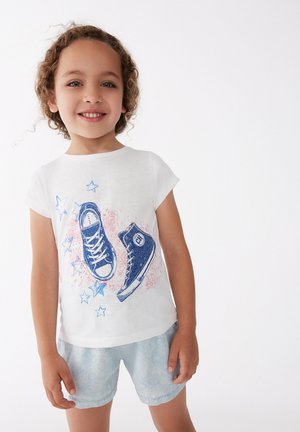Camiseta blanca de manga corta con un gráfico azul de dos zapatillas y estrellas. El niño lleva pantalones cortos de un patrón claro, mostrando un diseño juguetón.