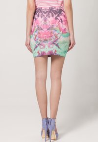 Femme portant une mini-jupe à imprimé tropical coloré et des chaussures à talons lavande, debout de dos face à la caméra.